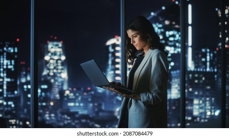 Big City Modern Office at Night: Successful Young Businesswoman Standing and Using Laptop. Beautiful Female Digital Entrepreneur Thinking of Investment Strategy for e-Commerce Project.