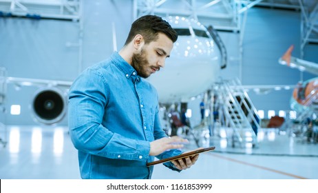 In Big Company Hangar Aircraft Maintenance Engineer Uses Tablet Computer while Standing Near Big New Shiny White Plane. - Powered by Shutterstock