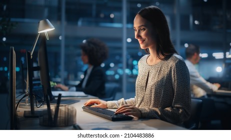 In Big Corporate Office at Night: Portrait of Confident Female Manager Using Computer, Businesspeople and Experts Working Around Her, Analysing Statistics, Commerce Data, Marketing Plans.