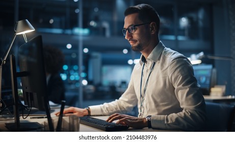 In Big Corporate Office At Night: Portrait Of Confident Manager In White Shirt Using Computer, Businesspeople And Experts Working Around Him, Analysing Statistics, Commerce Data, Marketing Plans.