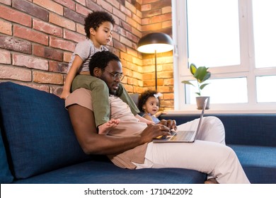 Big Daddy With Children Sitting At Home On The Couch Working Remotely On A Laptop