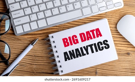 Big data analytics text on a spiral notepad with keyboard, mouse, glasses and pen on wooden desk . 