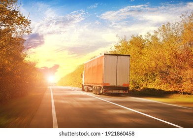 A Big Driving Fast Truck With A Red Trailer And Other Cars On A Countryside Road With Autumn Trees Against A Blue Sky With A Sunset