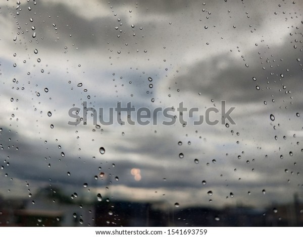 Big drops of rain on the window glass, during the storm.\
View of the overcast clouds and green trees and buildings. Urban\
view of rain drops falls on a window during a stormy day. Rain drop\
on window 