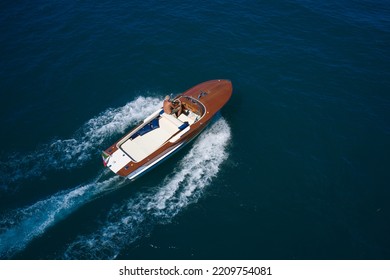 Big expensive wooden open boat with a man moving fast on dark blue water top view. Italian wooden speedboat moving up aerial view. Wooden expensive boat fast movement on dark water top view.