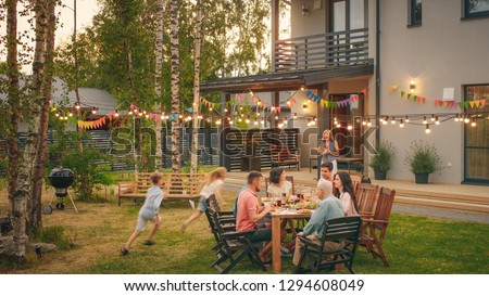 Big Family Garden Party Celebration, Gathered Together at the Table Family, Friends and Children. People are Drinking, Passing Dishes, Joking and Having Fun. Kids Run Around Table.