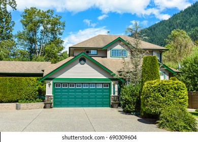 Big Family House With Wide Concrete Driveway, Double Wide Garage Door, Decorated Front Yard. Residential House With Mountain And Blue Sky Background