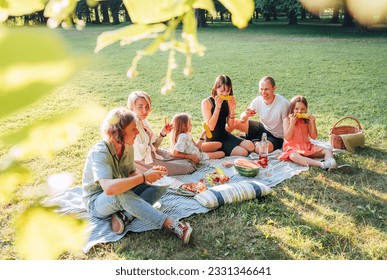 Big family sitting on the picnic blanket in city park during weekend Sunday sunny day. They are smiling, laughing and eating boiled corn and watermelon. Family values and outdoors activities concept. - Powered by Shutterstock