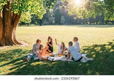 Big family sitting on picnic blanket in city park linden treeb during weekend Sunday sunny day. They are chatting and eating boiled corn and watermelon. Family values and outdoors activities concept - Powered by Shutterstock