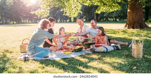 Big family sitting on the picnic blanket in city park during weekend sunny day. They smiling, laughing and eating watermelon, boiled corn,pie with cold tea. Family values, outdoors activities concept. - Powered by Shutterstock