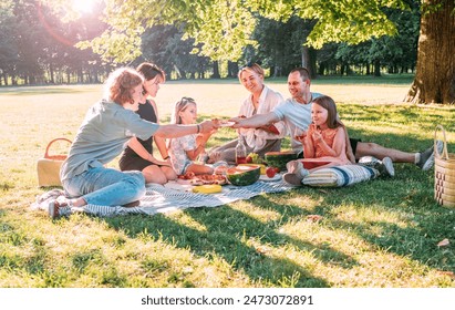 Big family sitting on the picnic blanket in city park during weekend sunny day. They smiling, laughing and eating watermelon, boiled corn,pie with cold tea. Family values, outdoors activities concept. - Powered by Shutterstock