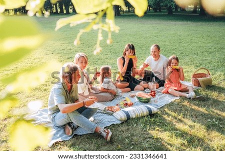 Big family sitting on the picnic blanket in city park during weekend Sunday sunny day. They are smiling, laughing and eating boiled corn and watermelon. Family values and outdoors activities concept.