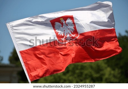 Big flag of Poland with national Polish emblem on it waves against green tree and blue sky