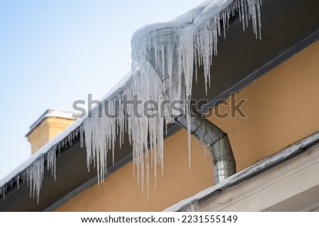 Big frozen icicles dangerously hanging from building edge on cold winter day, dangerous ice formation on metal house roof during bright sunny, but sub-freezing weather outside. Ice dam prevention