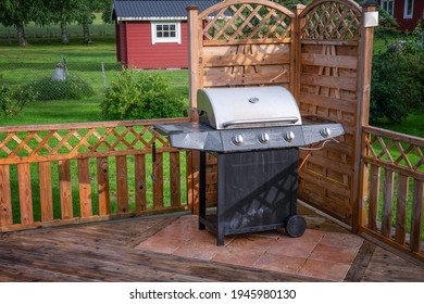 Big Gas Barbeque Grill, Stainless Construction With Black Bottom Stands At The Wooden Terrace Corner. Water Drops After Heavy Rain. Small Red Wooden House In Green Garden At Background. Summer Day