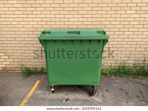 Big green commercial rubbish bin on side of\
building. Space to add text around the plastic skip, brick wall,\
car park floor, for background use. Waste management industry,\
removal business concept.