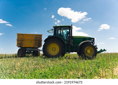 Big Green Tractor Standing In The Middle Of The Field With A Trailer. Outdoor Full-length Shot. Beautiful Weather, Blue Sky With White Clouds. High Quality Photo