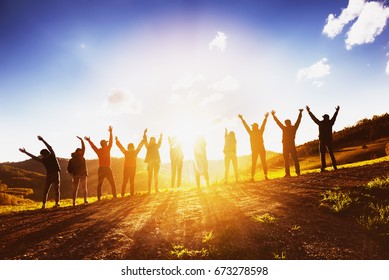Stor gruppe lykkelige venner står på solnedgang bakteppe med hevede armer sammen. Vennskap eller samarbeid konsept