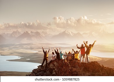 Большая группа людей, веселившихся в успех позируют с поднятыми руками на вершине горы против заката озер и гор. Концепция путешествий, приключений или экспедиции