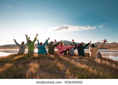 Big group of tourists sits with raised arms at sunset lake