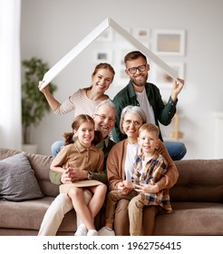 Big happy family, grandparents, mother, father with little kids son and daughter celebrating relocation in new home, sitting on the sofa under paper roof and smiling at camera. Mortgage loan concept