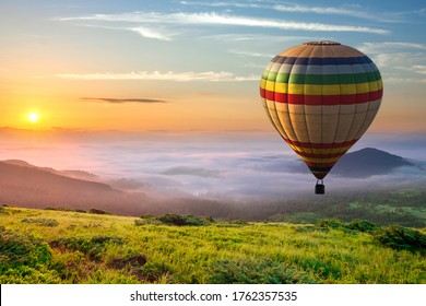 Big Hot Air Baloon Over Idyllic Landscape With Green Grass Covered Morning Mountains With Distant Peaks And Wide Valley Full Of Thick White Cloudy Fog.