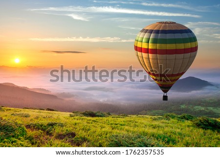 Big hot air baloon over idyllic landscape with green grass covered morning mountains with distant peaks and wide valley full of thick white cloudy fog.