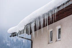 Big Icicles And Snow Hanging Over The Rain Gutter On A Roof Of A Traditional Wooden House In The Mountains In Winter Could Be Dangerous. Trees At The Background.