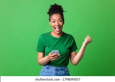 Big Luck. Emotional African Girl Holding Cell Phone Shouting And Gesturing Yes Standing On Green Wall