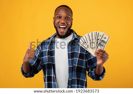 Big Luck. Joyful Black Guy Holding Money Cash Gesturing Thumbs Up Posing Standing Over Yellow Studio Background. Profit And Financial Success, Rich African Man Showing Like Gesture