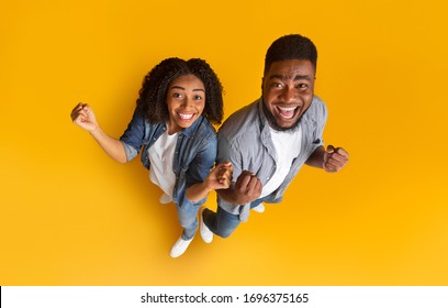 Big Luck. Top view of excited black couple celebrating success or victory with raised hands, clenching fists while standing on yellow background
