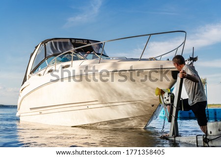 Big luxury cabin motorboat cruiser yacht launching at trailer ramp on river or lake. Mechanic worker man assist putting boat in calm water surface. Luxury rich fishing leisure recreation lifestyle