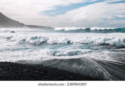 Big and massive atlantic ocean waves rolling to the coast with black sand beach. Huge ocean waves with crystal blue water on high tide with cliffs, rocks and dramatic blue sky on background.