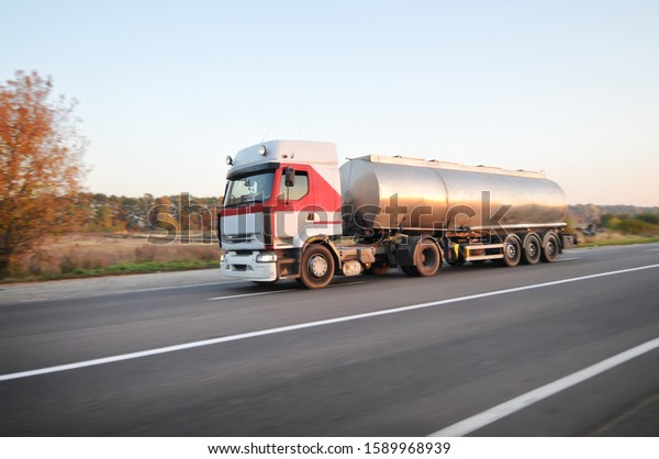 Big metal fuel\
tanker truck shipping fuel on the countryside road in motion with\
forest against clear sky