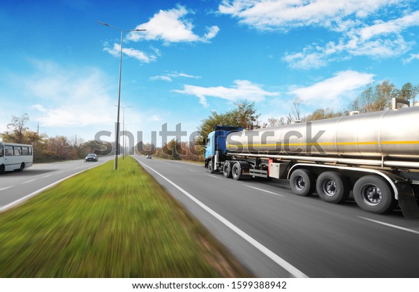 Big metal fuel tanker truck shipping fuel with\
other cars on the countryside road in motion with trees against\
blue sky with clouds