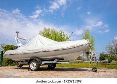 Big Modern Inflatable Motorboat Ship Covered With Grey Or White Protection Tarp Standing On Steel Semi Trailer At Home Backyard On Bright Sunny Day With Blue Sky On Background. Boat Vessel Storage