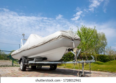 Big modern inflatable motorboat ship covered with grey or white protection tarp standing on steel semi trailer at home backyard on bright sunny day with blue sky on background. Boat vessel storage