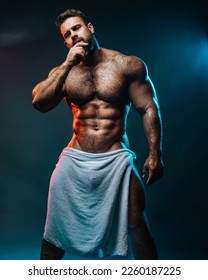 Big muscled man in white towel. Handsome hunk posing is studio. Naked male model with muscles and hairy body at dark background. Bodybuilder wearing only bath towel. Sexy athlete with six pack abs.
