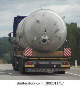 Big New Barrel On A Low-frame Trawl Semi Truck Drive On Suburban Asphalt Road Close-up Back, Oversized Cargo Transportation Logistics On Lowbed Long Vehicle On Summer Day