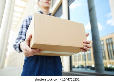 Big Packed Carton Box In Hands Of Delivery Man In Workwear Walking Along Modern Building