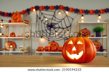 Big pumpkin with carved funny glowing face standing on background of Halloween decor. Close up of jack-o-lantern standing on right side on wooden table. Halloween holiday concept. Blurred background
