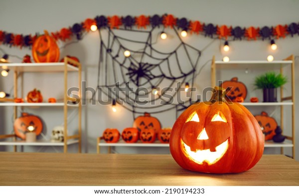 Big pumpkin with carved funny glowing face\
standing on background of Halloween decor. Close up of\
jack-o-lantern standing on right side on wooden table. Halloween\
holiday concept. Blurred\
background