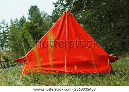 Big red camping tent constructed for relaxation on green lawn near forest in non-urban place. Charming bright shelter pitched on grass in summer day, attached with ropes and ready for night.