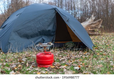 Big red gas burner near a tourist tent on a background of autumn forest on cloudy day, adventure theme. Camping equipment for making food