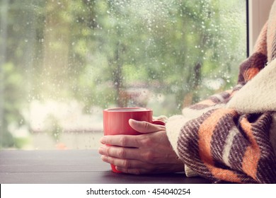 Big Red Mug In The Hands Thrown Over With A Warm Blanket Against The Window With Rain Drops / Warming Drink At Home