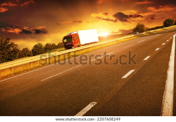 Big red truck with a\
white trailer on a counstryside road with trees against an evening\
sky with a sunset