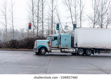 Big Rig American Classic Long Haul Semi Truck With Dry Van Semi Trailer Turning On Traffic Light Crossroad Intersection Wet Road With Raining And Foggy Weather Going To Warehouse For Cargo Unloading