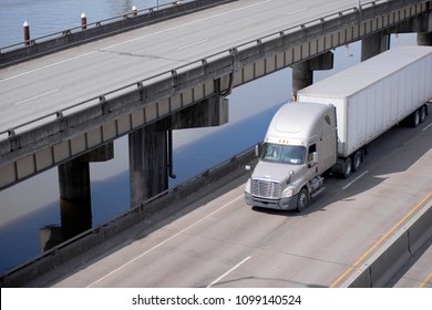 Big rig beige American bonnet long haul semi truck tractor transporting commercial cargo in dry van semi trailer running on overpass road along the river and upper level elevated highway
