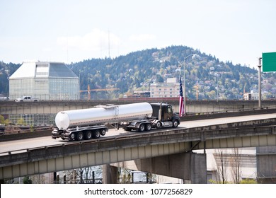 Big Rig Black Semi Truck Transporting Fuel In Long Round Stainless Steel Tank Semi Trailer Going On The City Overpass Road Intersection With American Flag And With Hill And Building On Background