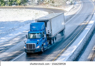 Big rig blue bonnet day cab semi truck with roof spoiler and refrigerator unit on the front wall of reefer semi trailer transporting commercial cargo driving on the turned winter wet road with snow